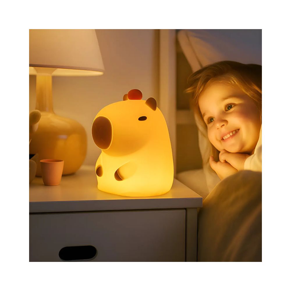 Fotografia em estilo clean e minimalista com fundo branco luminoso, apresentando uma luminária de mesa em formato de capivara acesa. A capivara é feita de silicone macio e emite uma luz quente e suave em tom amarelado. Ela está posicionada sobre uma superfície clara ao lado de um pequeno vaso de cerâmica branca com um ramo de eucalipto e um guardanapo de linho bege, transmitindo uma atmosfera calma, infantil e acolhedora.